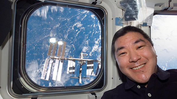 Daniel Tani smiles on the space shuttle with a view of the ISS in the window behind him
