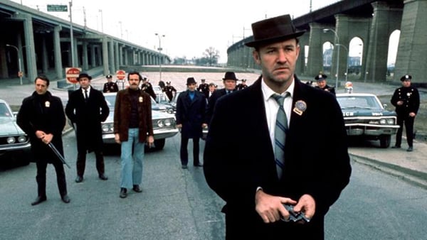 A still from "The French Connection" showing a man in a hat with a group of men standing several feet behind him