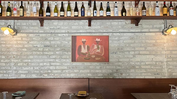 A brick wall with a painting, seats and tables below, and wine bottles on shelves above
