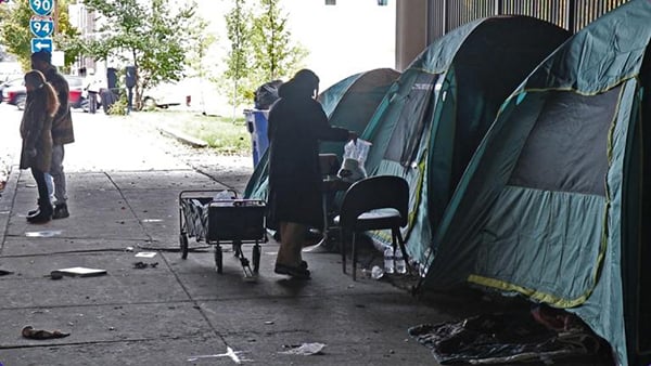 A person pulling a wagon delivers a care package to a tent under an overpass