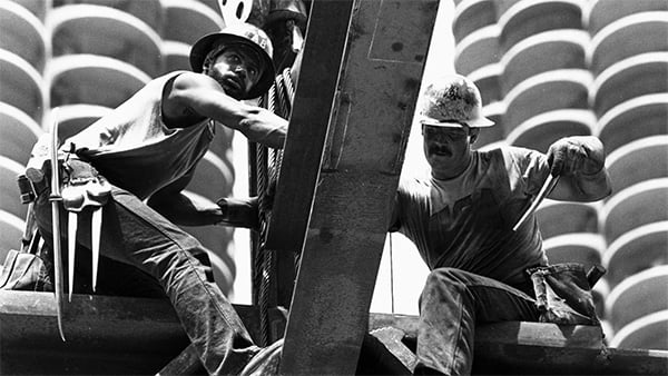 Two construction workers stand on steel girders with Marina City in the background