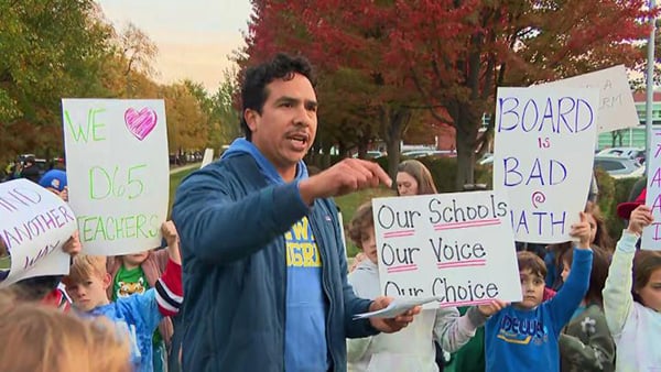 A man speaks to a crowd with a group of school kids holding protest signs behind him
