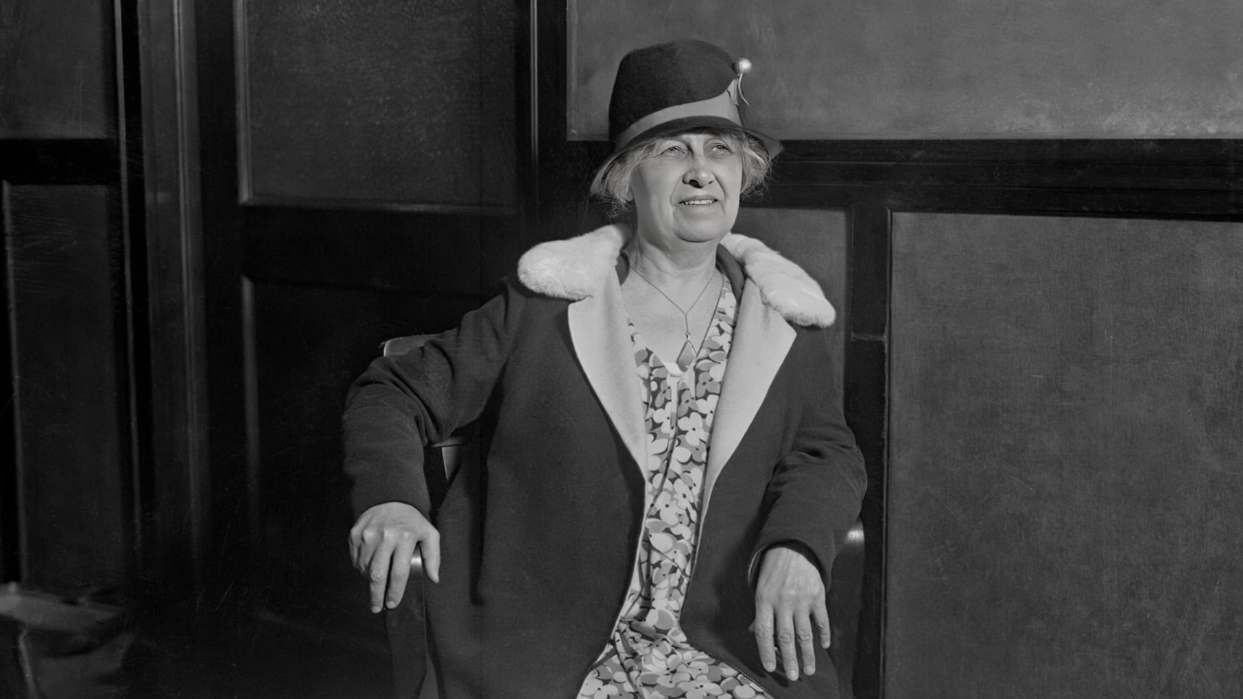 A black and white photo of a woman sitting in a chair in a coat and hat