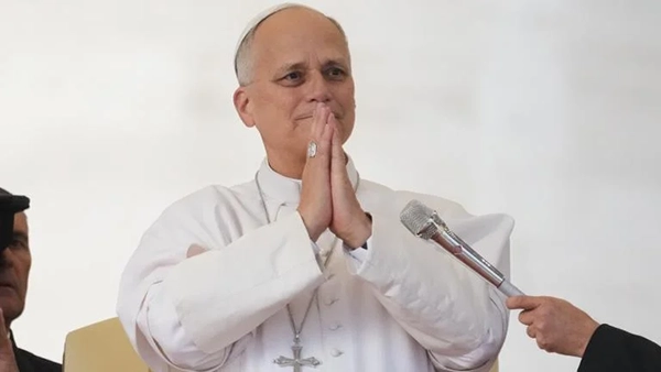Pope Leo holds his hands up in prayer while a hand off screen holds up a microphone to him