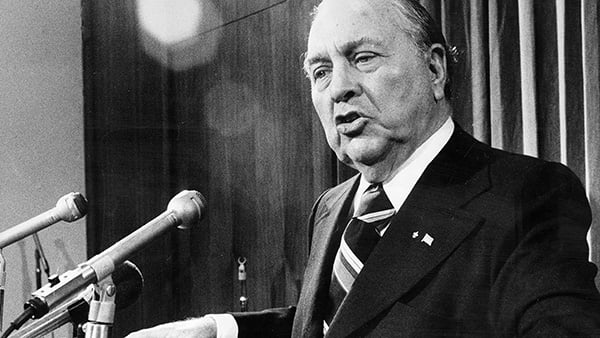 A black and white photo of Richard J. Daley in a suit standing at a podium behind microphones