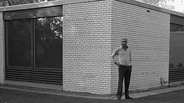 John Moutossamy stands at the corner of a brick building with glass windows in a black and white photo