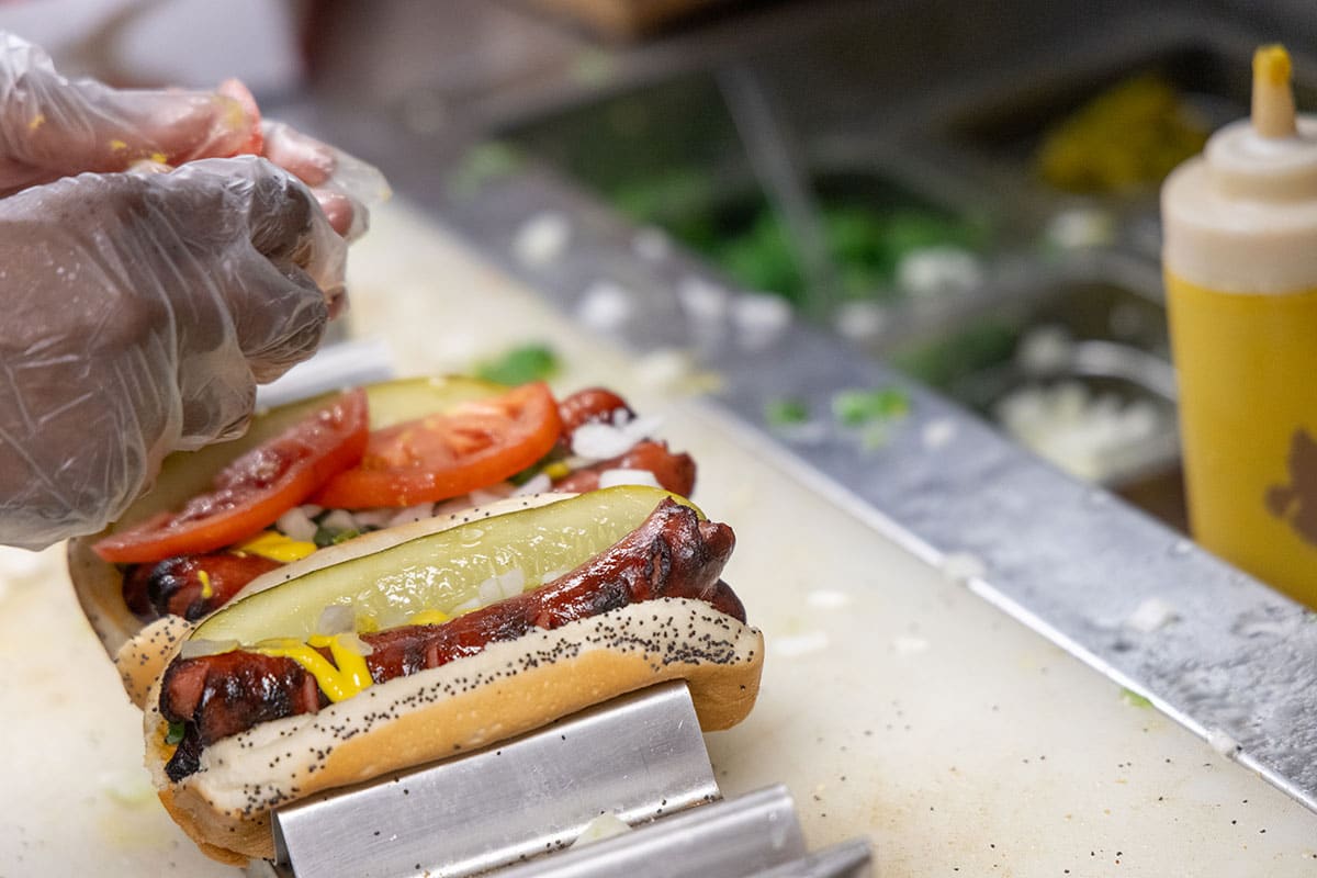 Closeup of a Chicago hotdog being topped with condiments behind the counter
