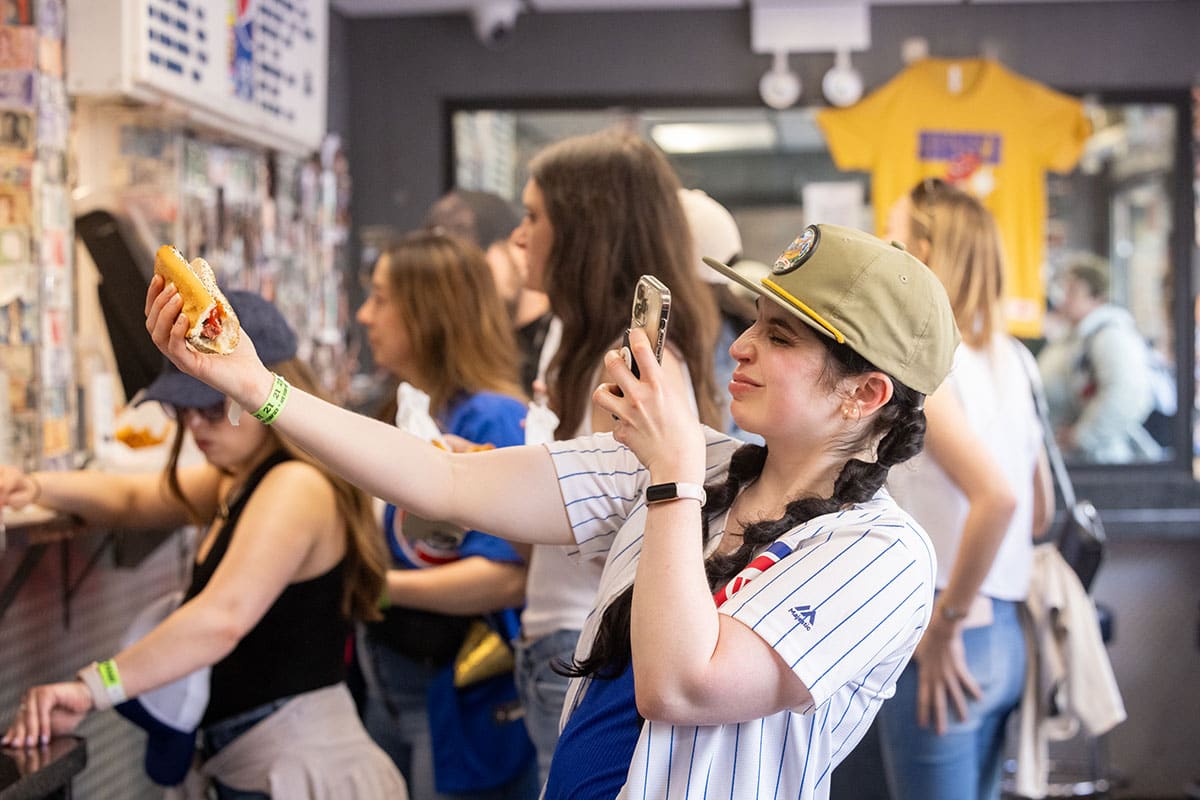 Woman holds her arm out with her hotdog while she takes a photo of it on her phone