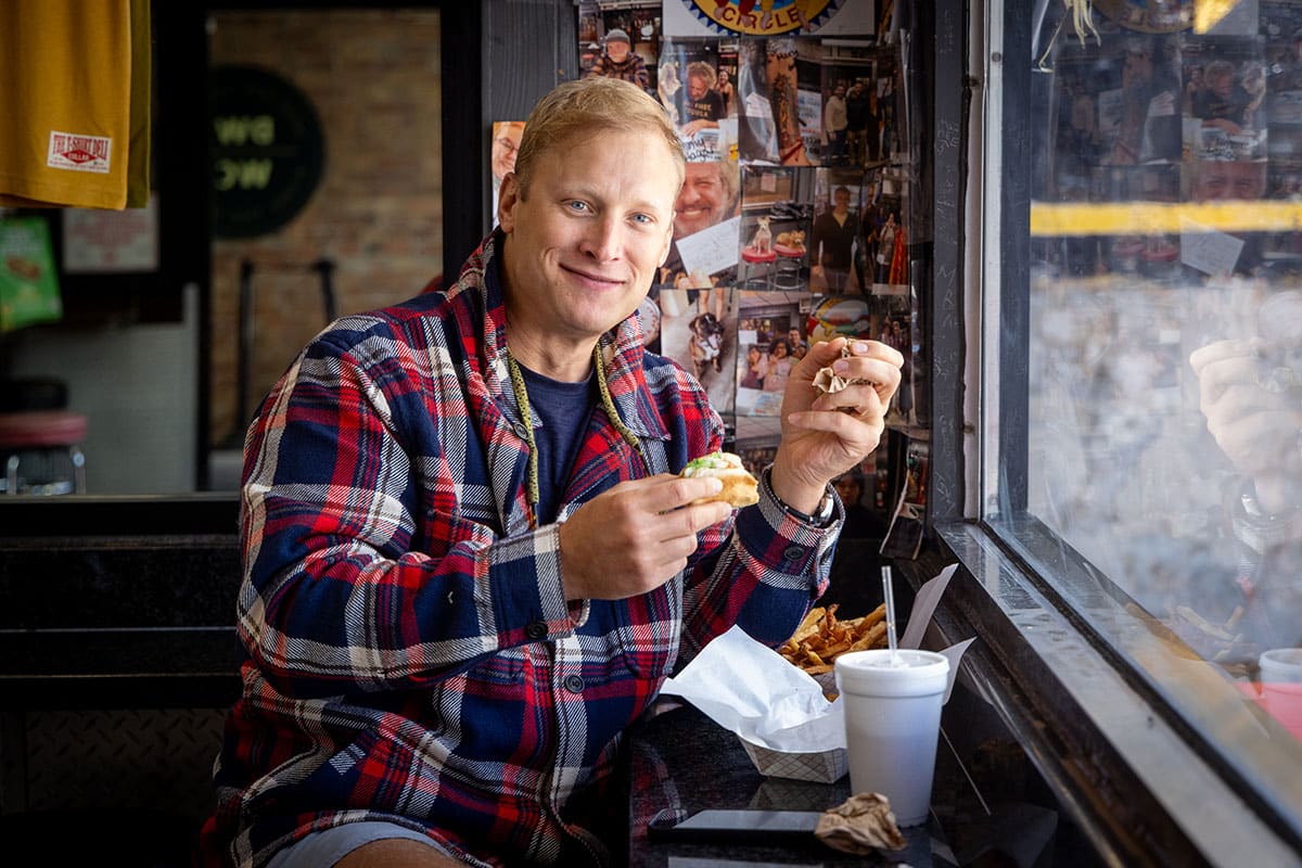 Gregory Piper smiles at the camera holding his half-eaten hotdog sitting at the counter