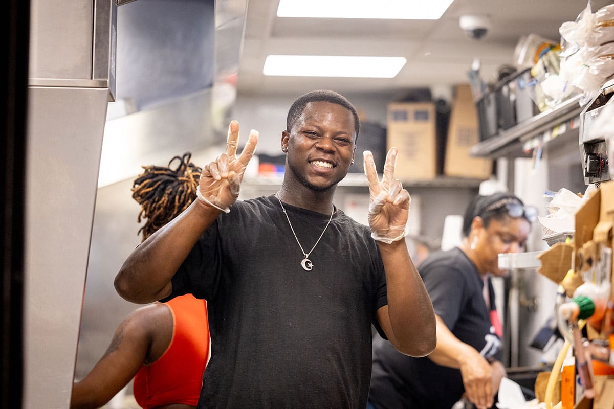 Joseph Taylor wearing plastic gloves raises two piece signs while working behind the counter