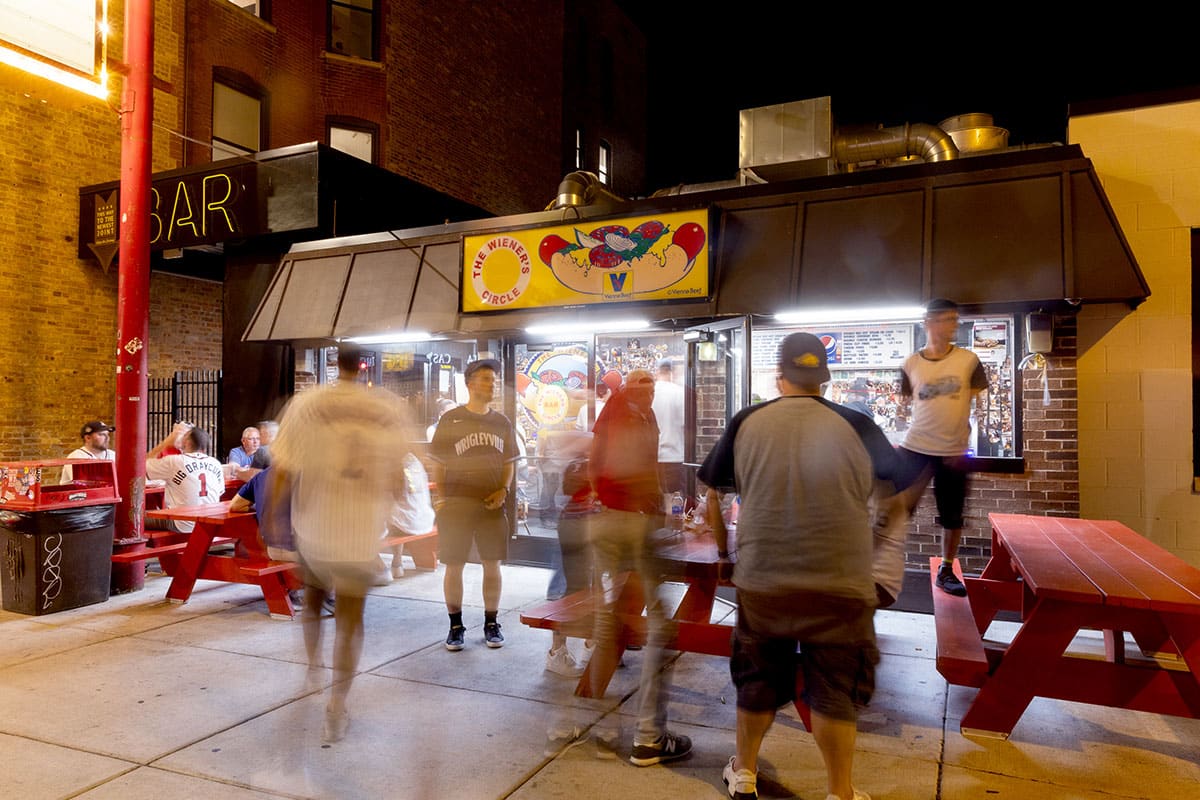 Nighttime view of the Weiner's Circle as people sit and stand around the red picnic tables
