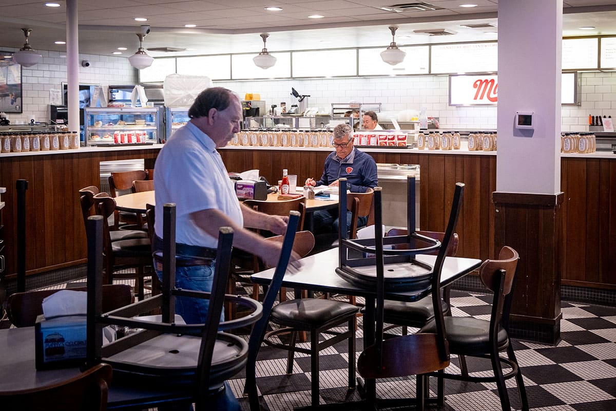 A restaurant setting with two men, one stacking chairs and another seated, dim lighting.