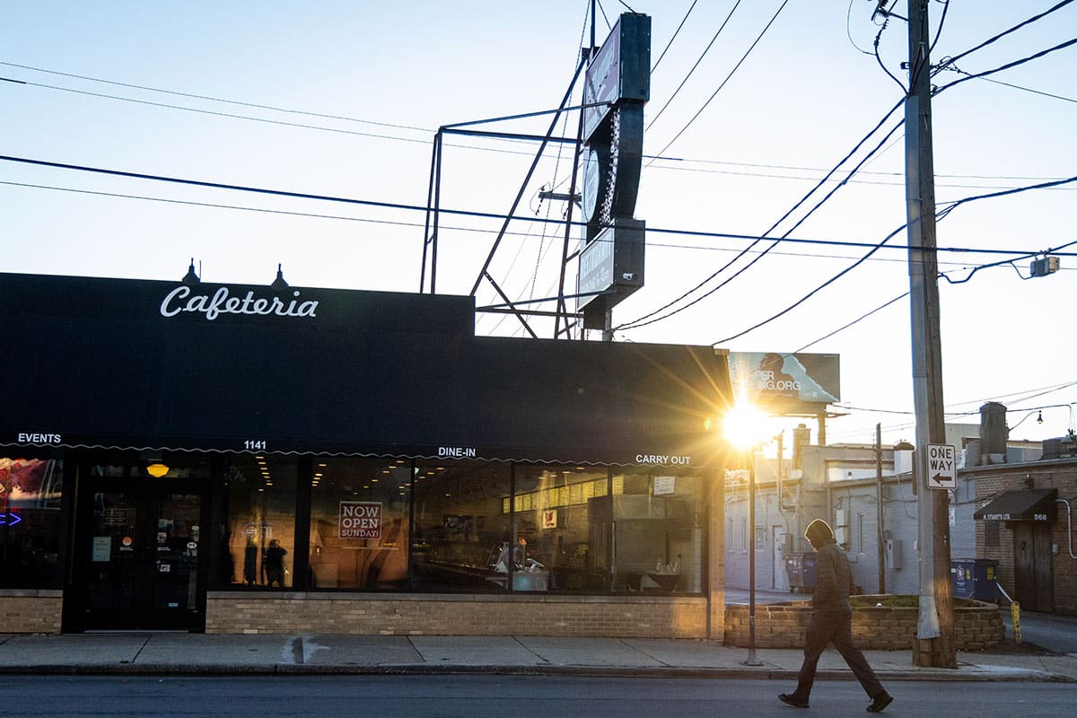 A person strolls towards Manny's Restaurant at sunrise, casting warm light.
