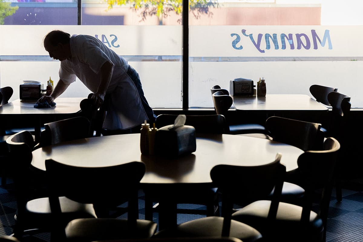 A restaurant worker cleans a table in a sunlit dining area with empty chairs.