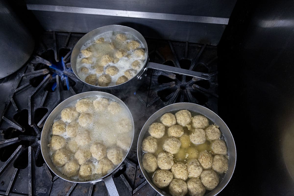 Three pots boiling matzo balls on a stove top, surrounded by steam and metal surfaces.