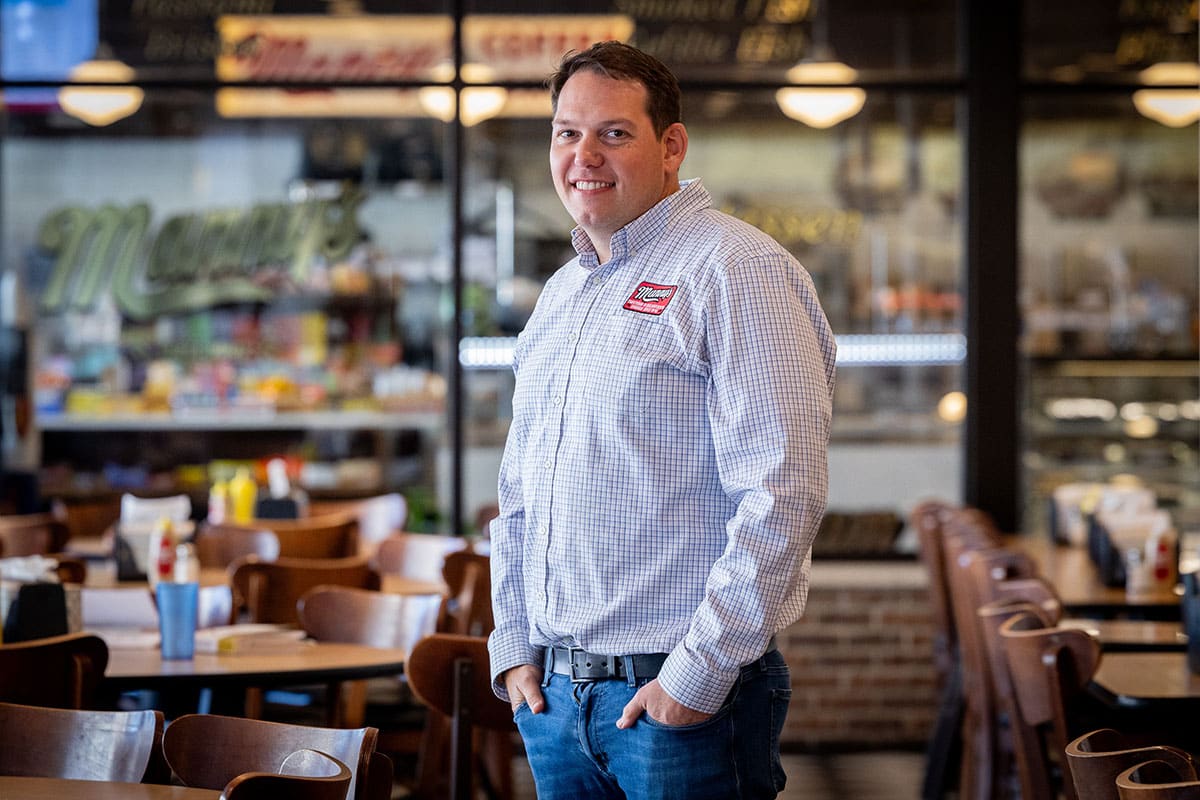 A man in a button-up shirt stands smiling in a casual restaurant setting.