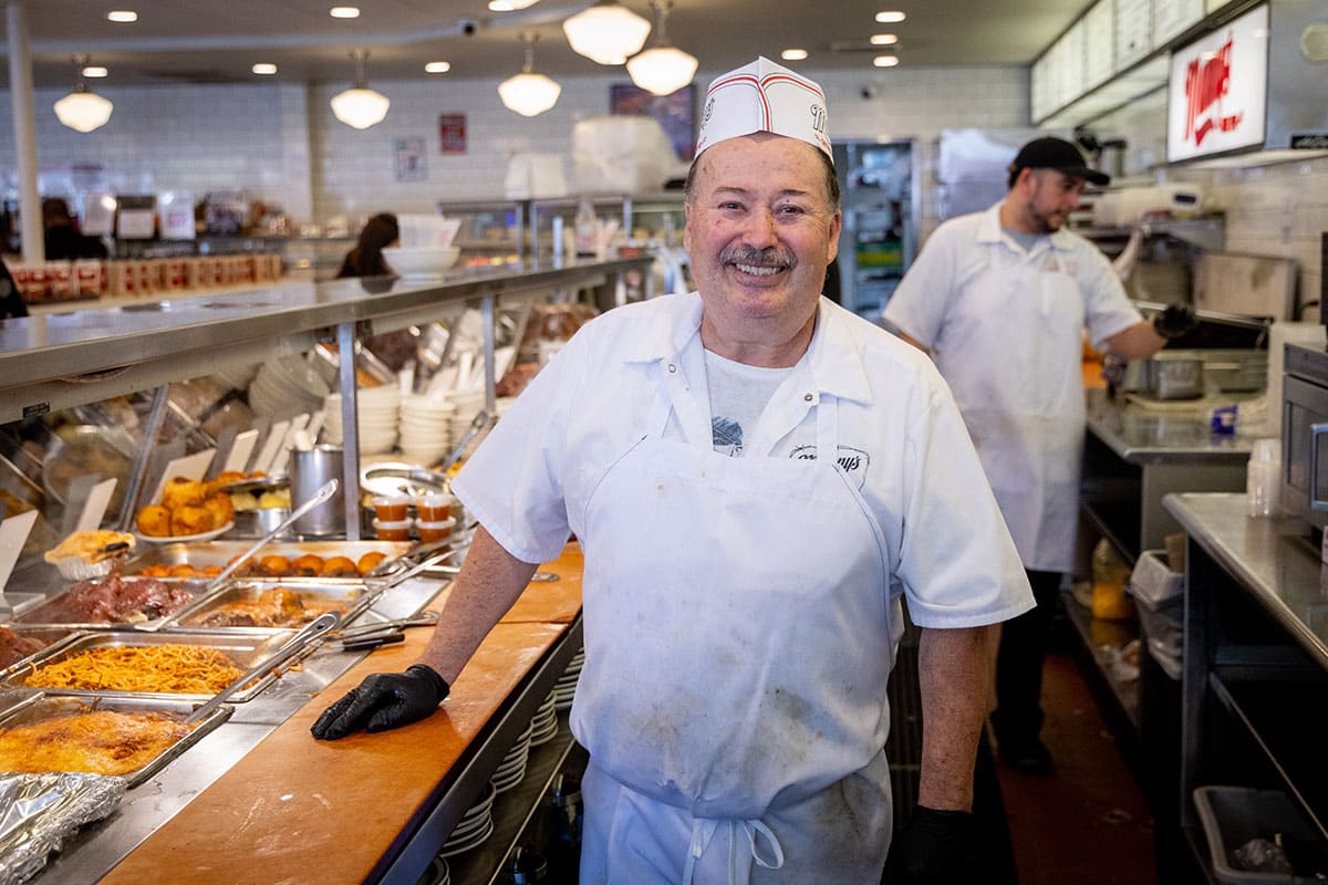 Smiling chef behind the deli counter, standing near food trays.