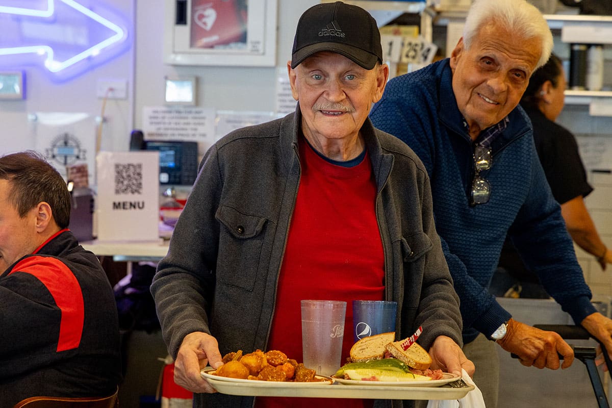 An elderly man holds a tray of food, smiling, while another man stands behind him.