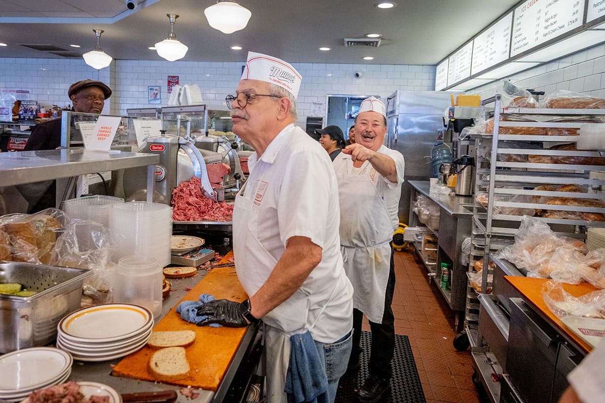 Busy deli counter with two workers in white uniforms, one smiling and pointing while the other prepares food.