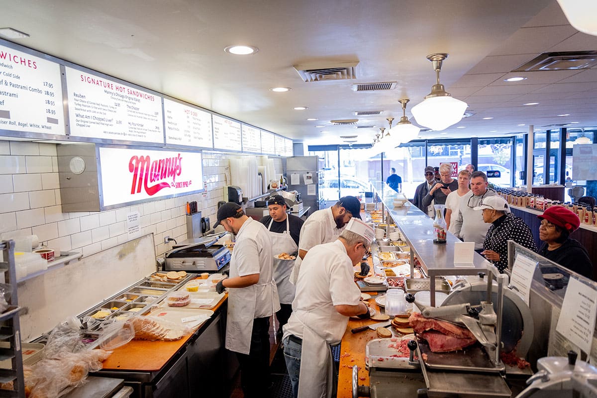 Busy deli interior with chefs preparing food and customers at the counter.