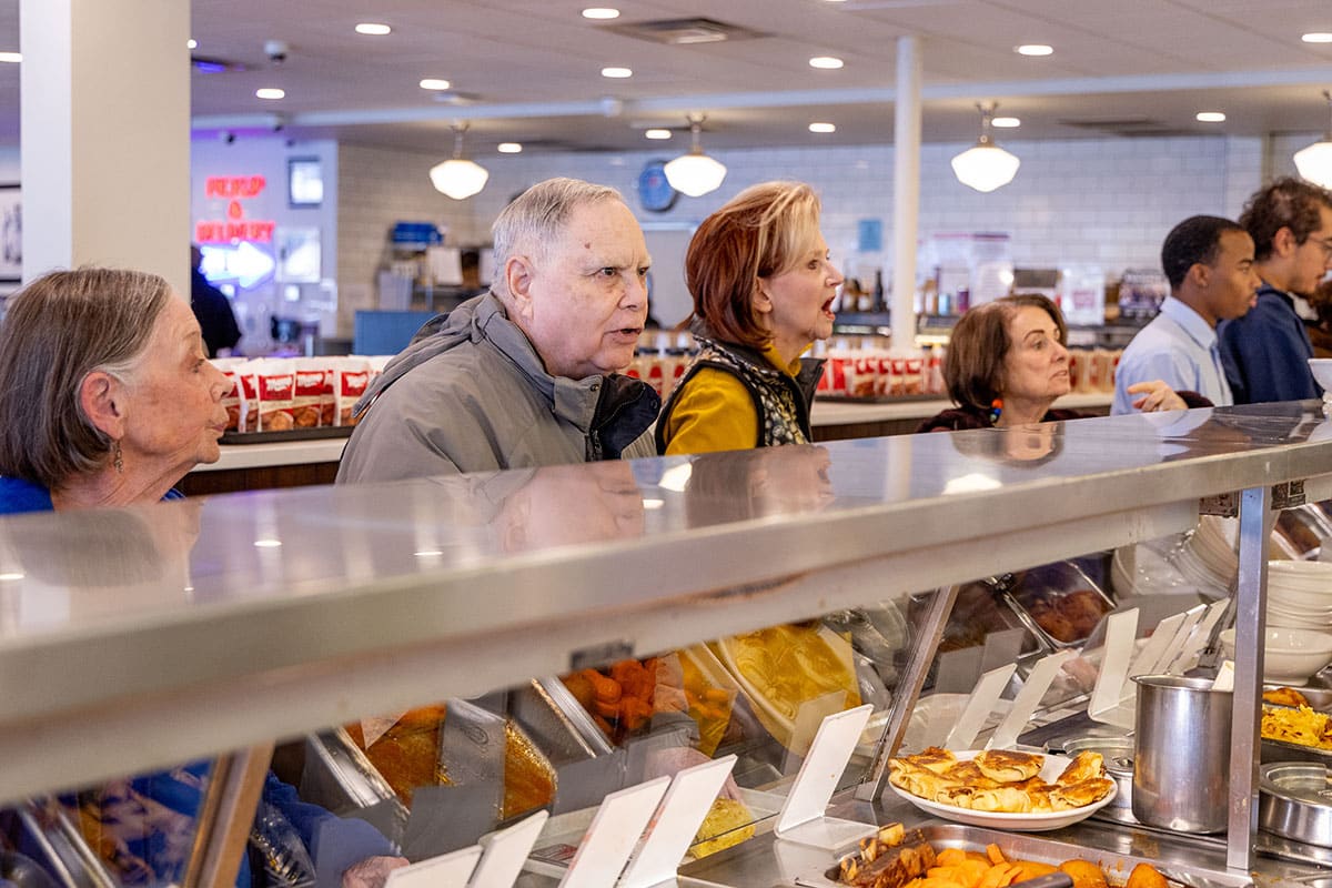 A group of people waiting in line and telling their orders at the deli counter.