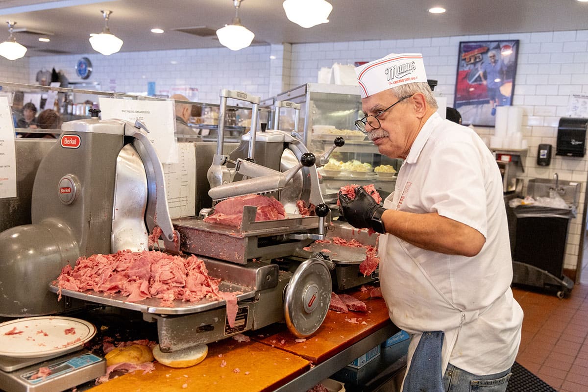 A butcher in a white apron and hat slices meat at a deli, surrounded by equipment and food.