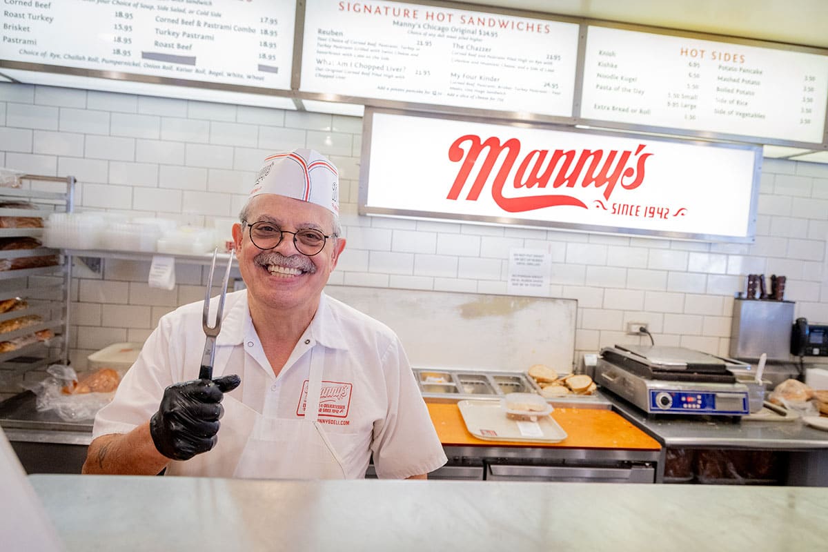 Smiling chef holding a meat fork in a busy deli with menu displayed in the background.