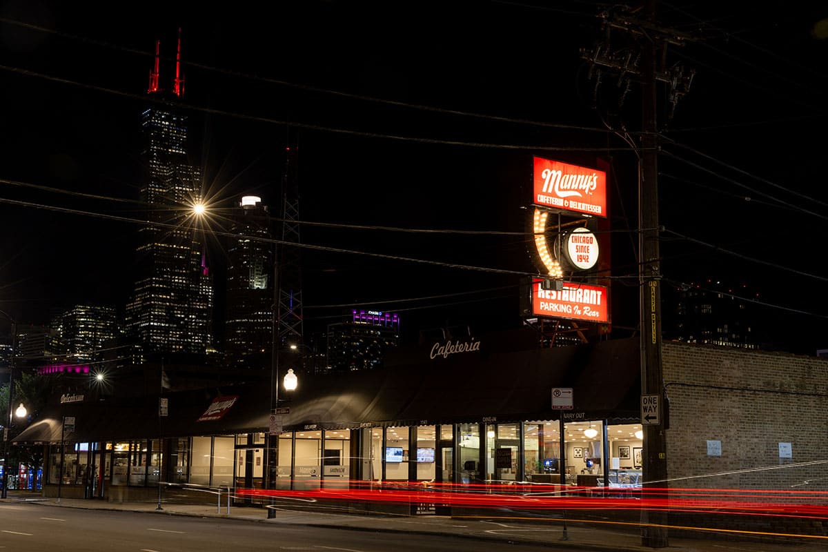 Night scene of a restaurant with city skyline and illuminated sign in the background.