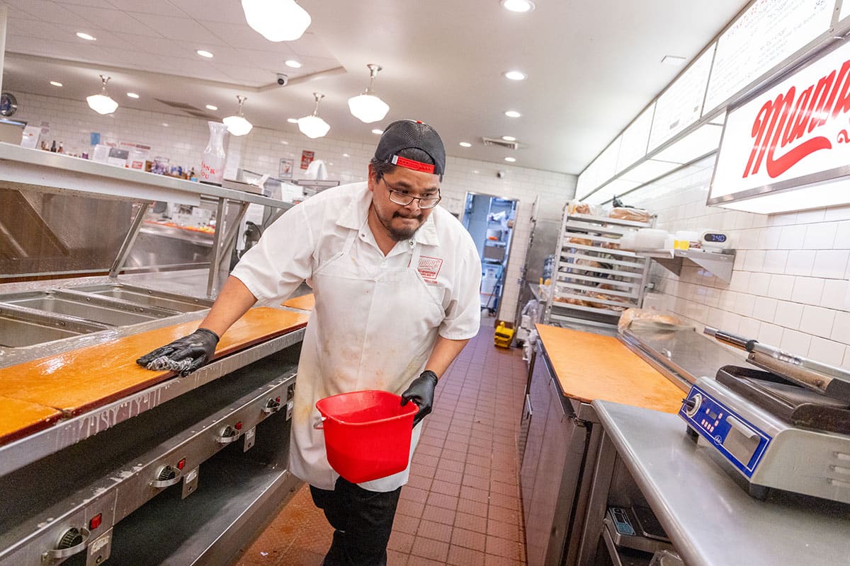 A restaurant worker in a white uniform cleans a countertop with a red bucket nearby.
