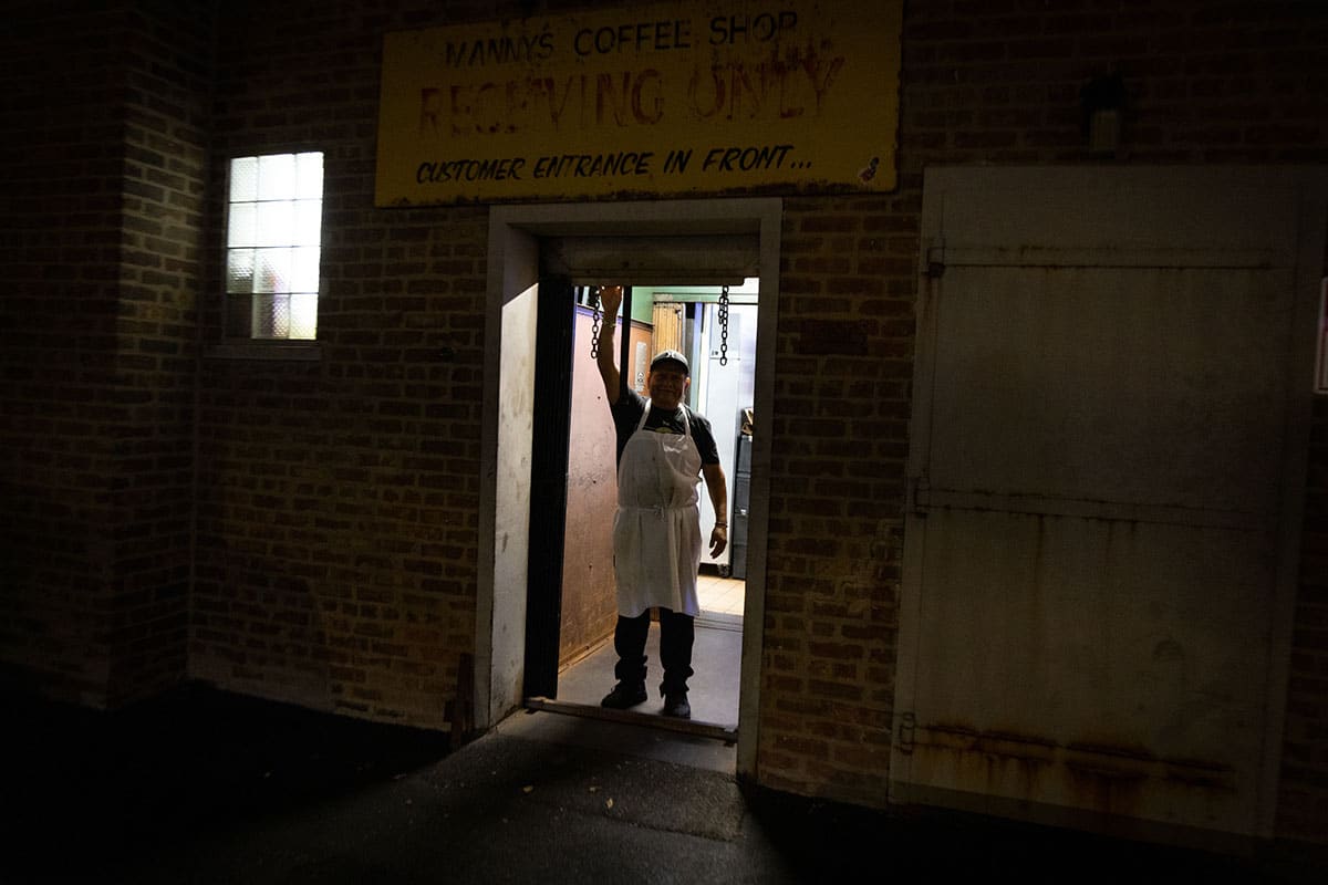 A person in an apron stands in a doorway at night with arm raised, in a dimly lit setting.