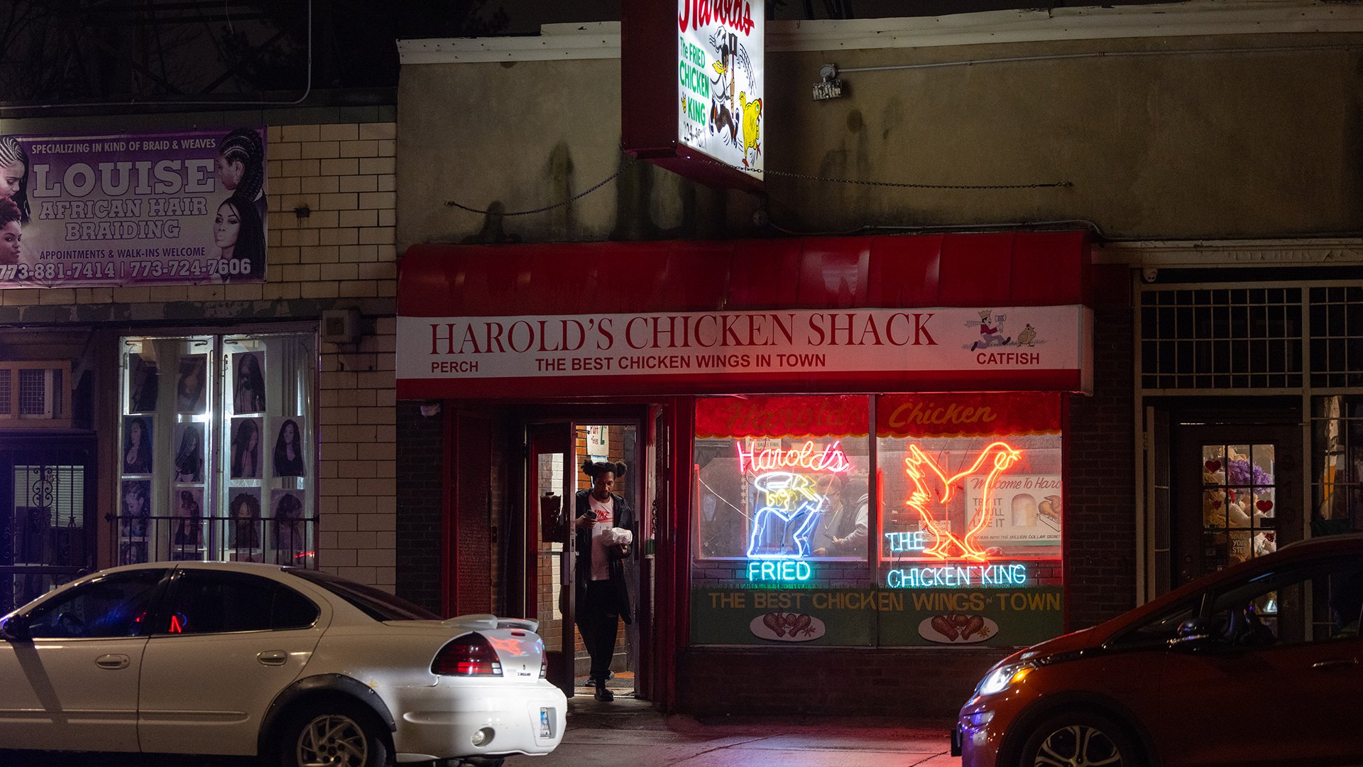 Harold's Chicken shown at night from outside with a neon sign in the window and a patron leaving