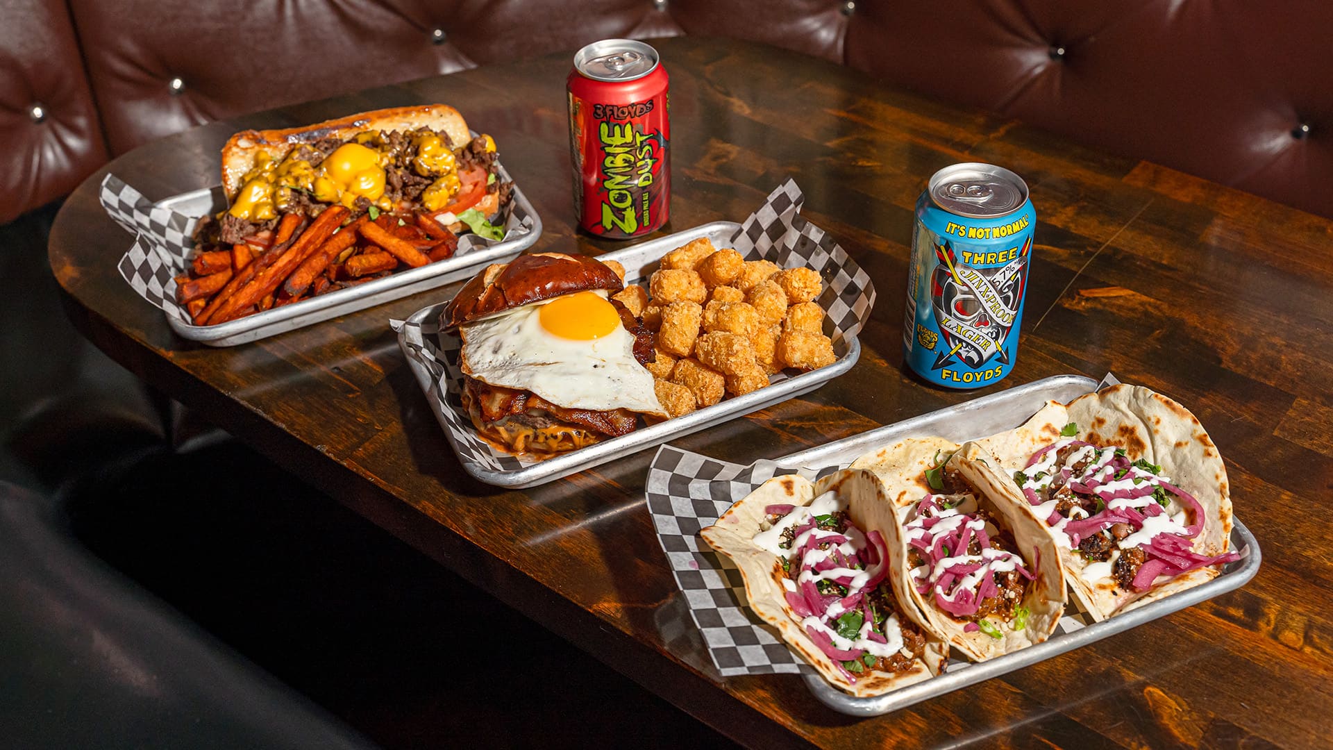 A restaurant booth with two cans of beer and three trays of food on the table.