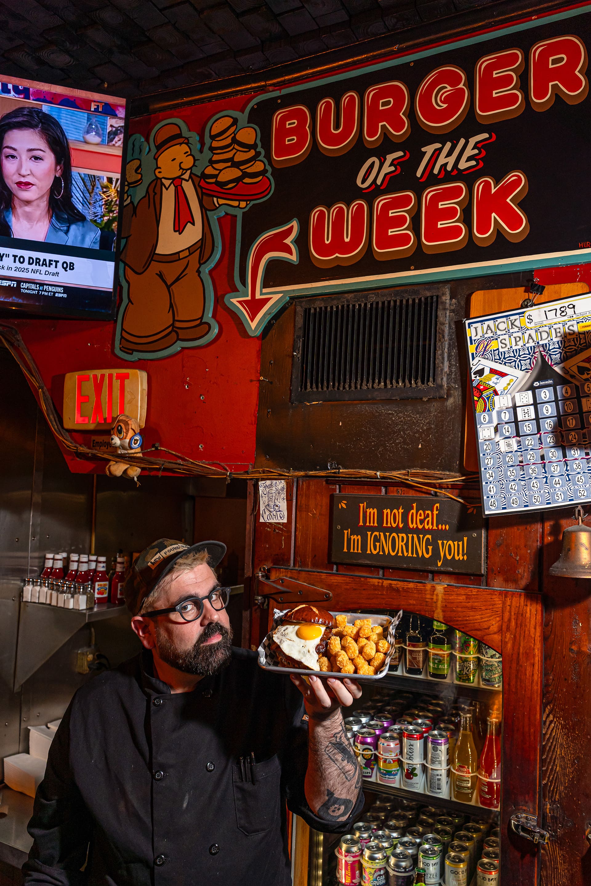 Larry Rosinko holds up a tray with tater-tots and a hamburger topped with a sunny-side-up egg under a 'burger of the week' sign.