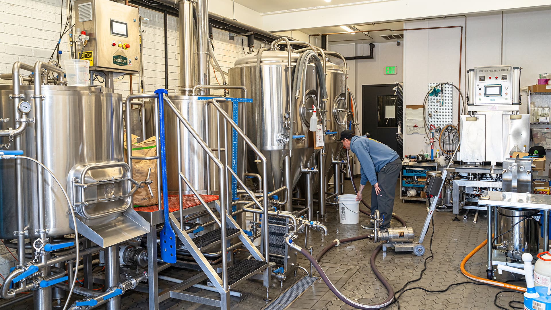 A worker places a bucket next to one of the large stainless steel brewing vessels