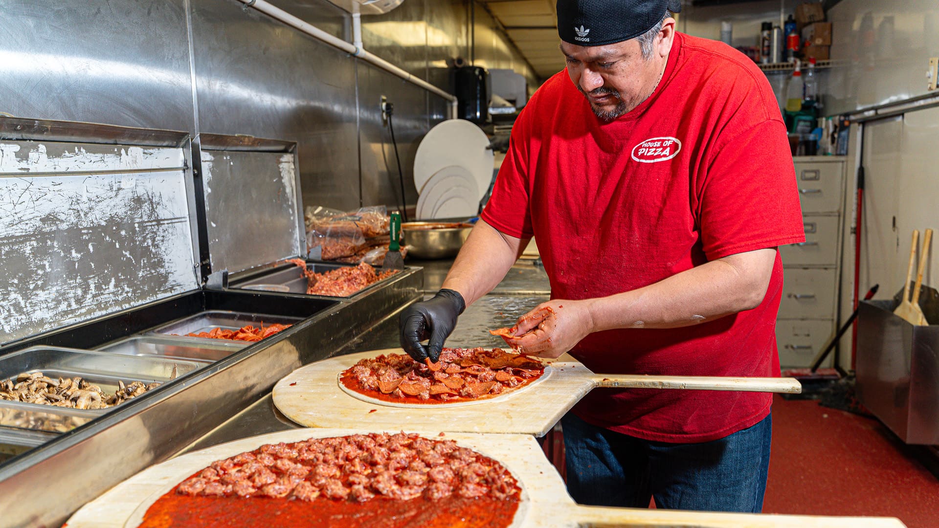 A chef tops a pizza with sausage while another sits in the forground with sauce and 1/2 sausage waiting for cheese to be added