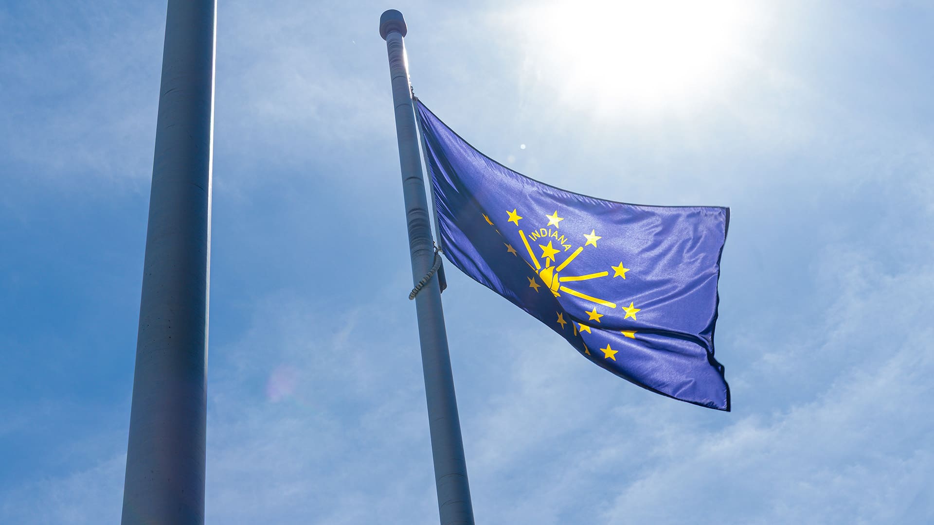 The Indiana state flag waves in the wind with the sun and clouds above.