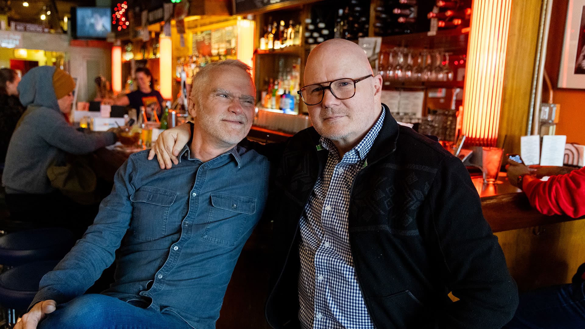 Two men at a bar, with one with their arm around the other, surrounded by a vibrant bar setting.