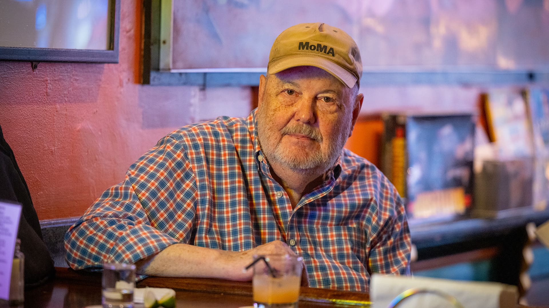 An older man in a hat sits at a bar, looking contemplative while enjoying his drink.