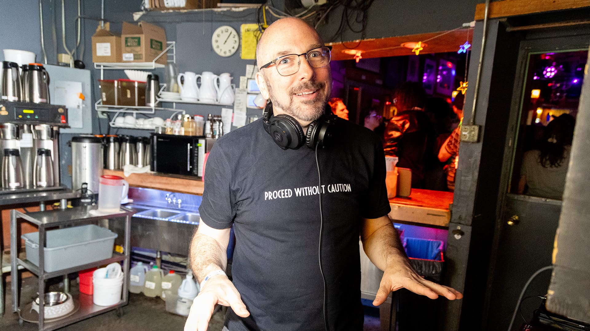 A man wearing headphones around his neck stands in a kitchen.