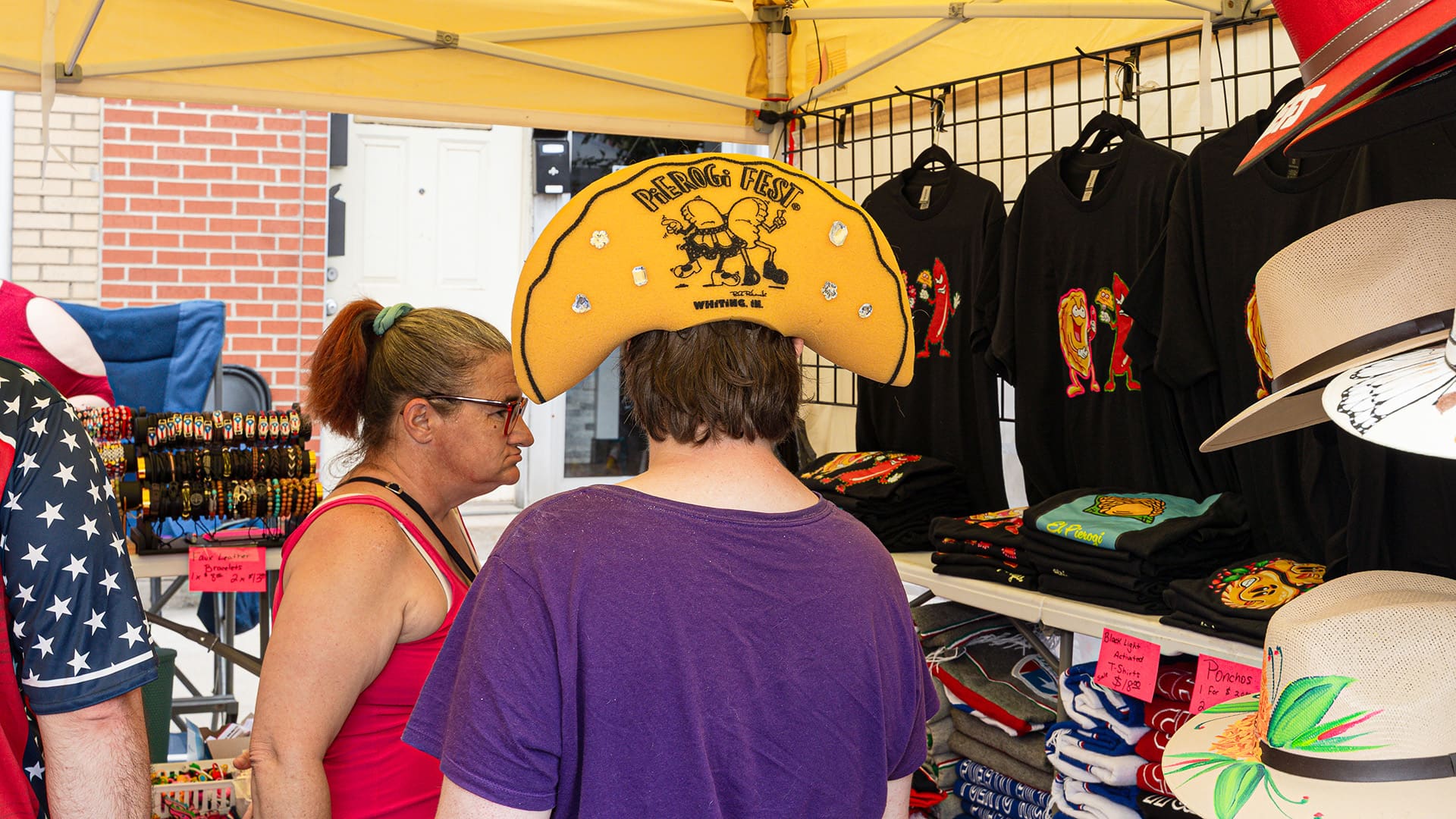 A person wears a Pierogi Fest foam hat, which is shapded like a pierogi while looking at wears in a vendors tent