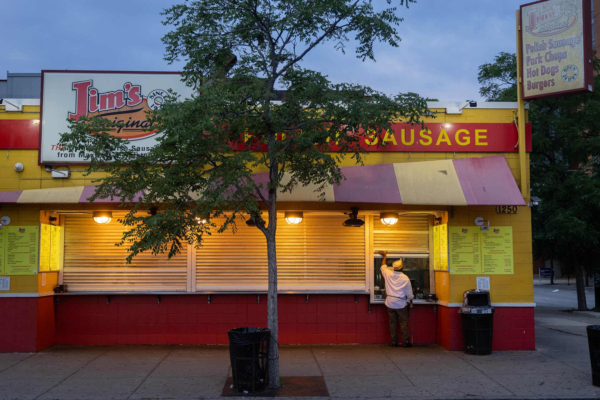 A worker opening the pickup window of Jim's Original at dawn, with the stand' shutters still down