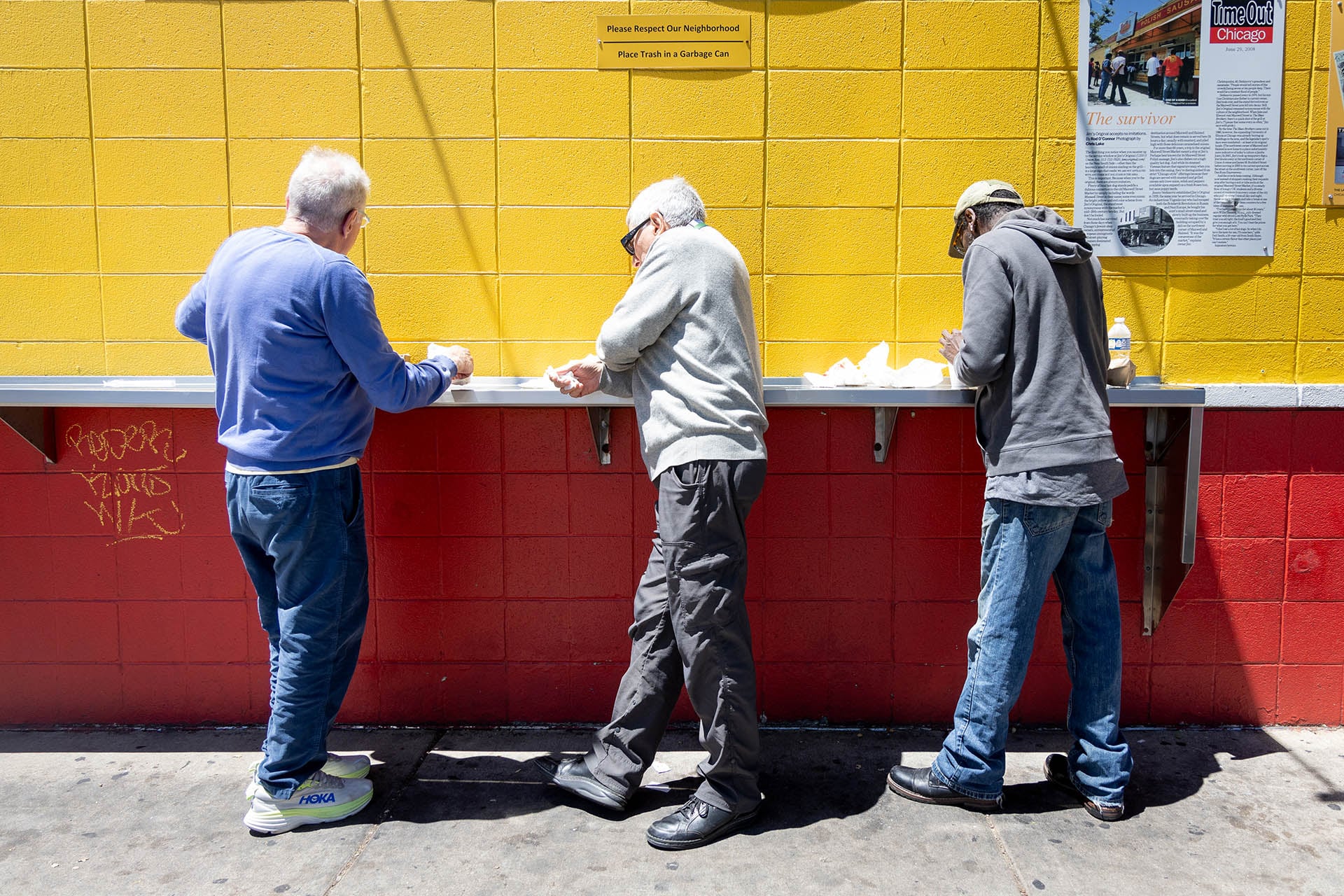 Three customers eating at the metal counter along the yellow and red exterior wall of Jim’s Original