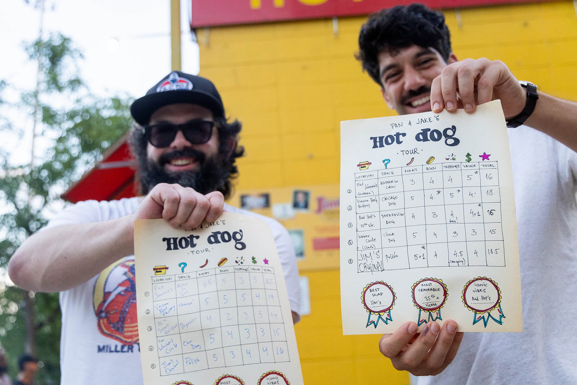 Pan Panayotov and Jake Nelson holding hand-drawn scorecards from their Chicago hot dog tour outside Jim’s Original