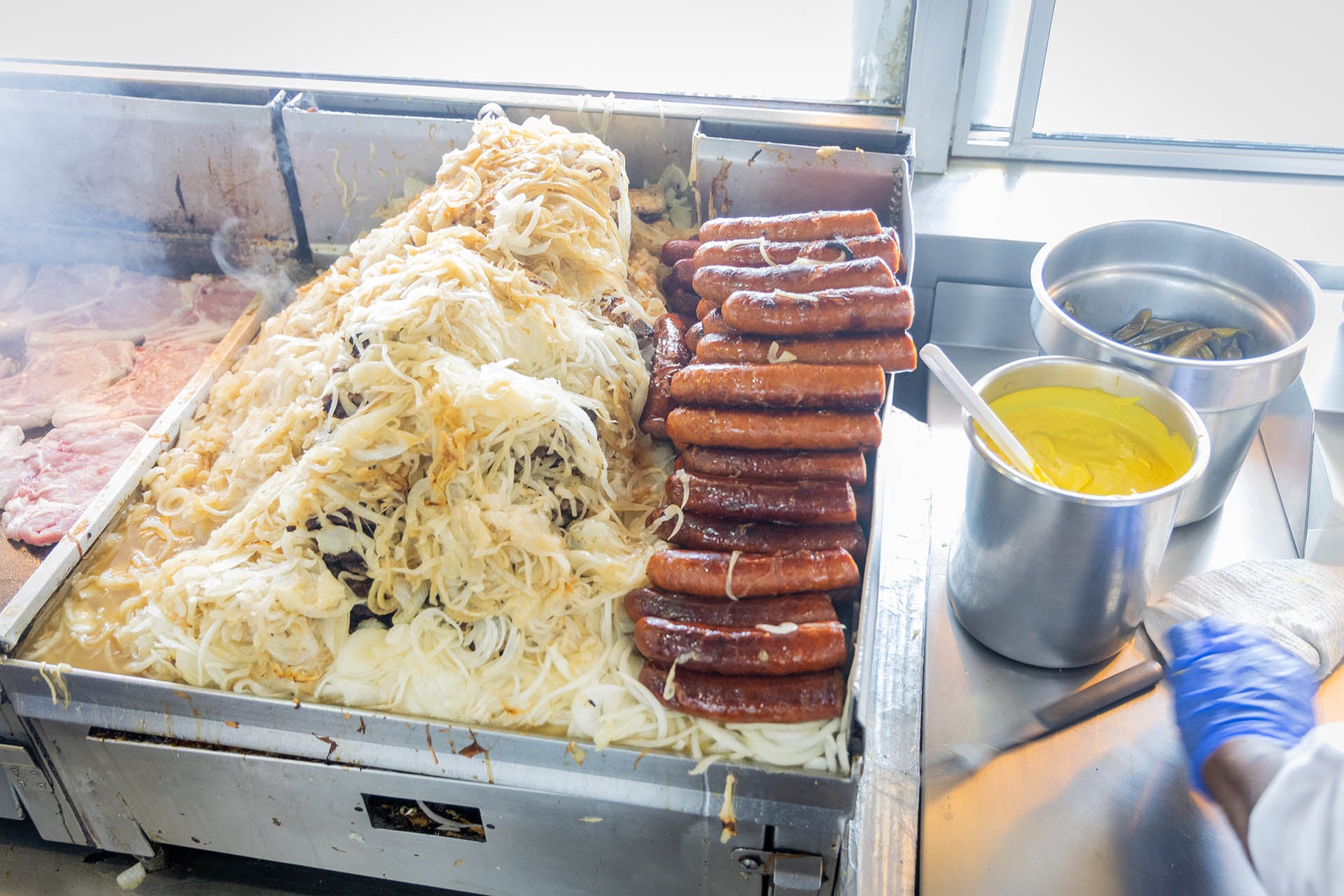 The griddle at Jim’s Original piled with Polish sausages and grilled onions, with pork chops cooking alongside and a container of yellow mustard nearby