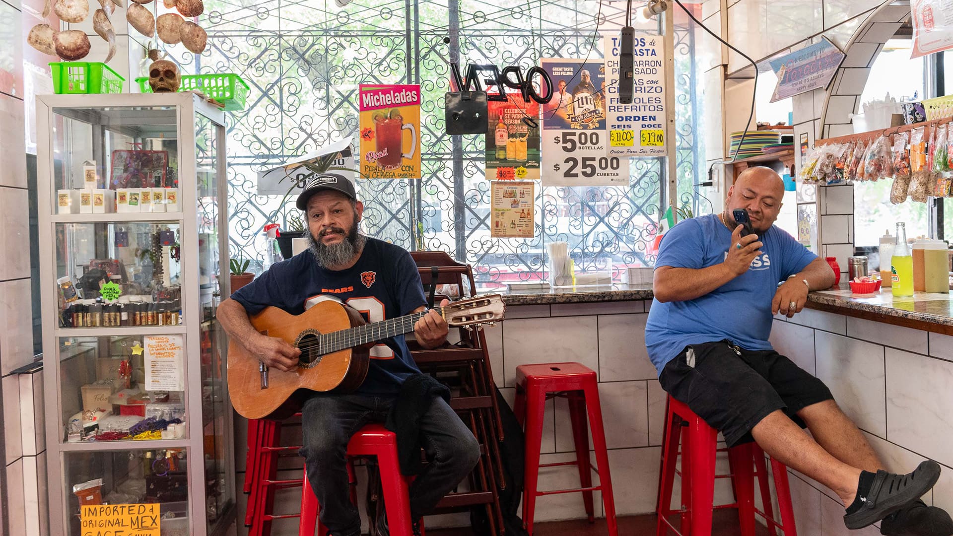 A man plays a guitar on a stool inside a store while another man sitting nearby films on his phone