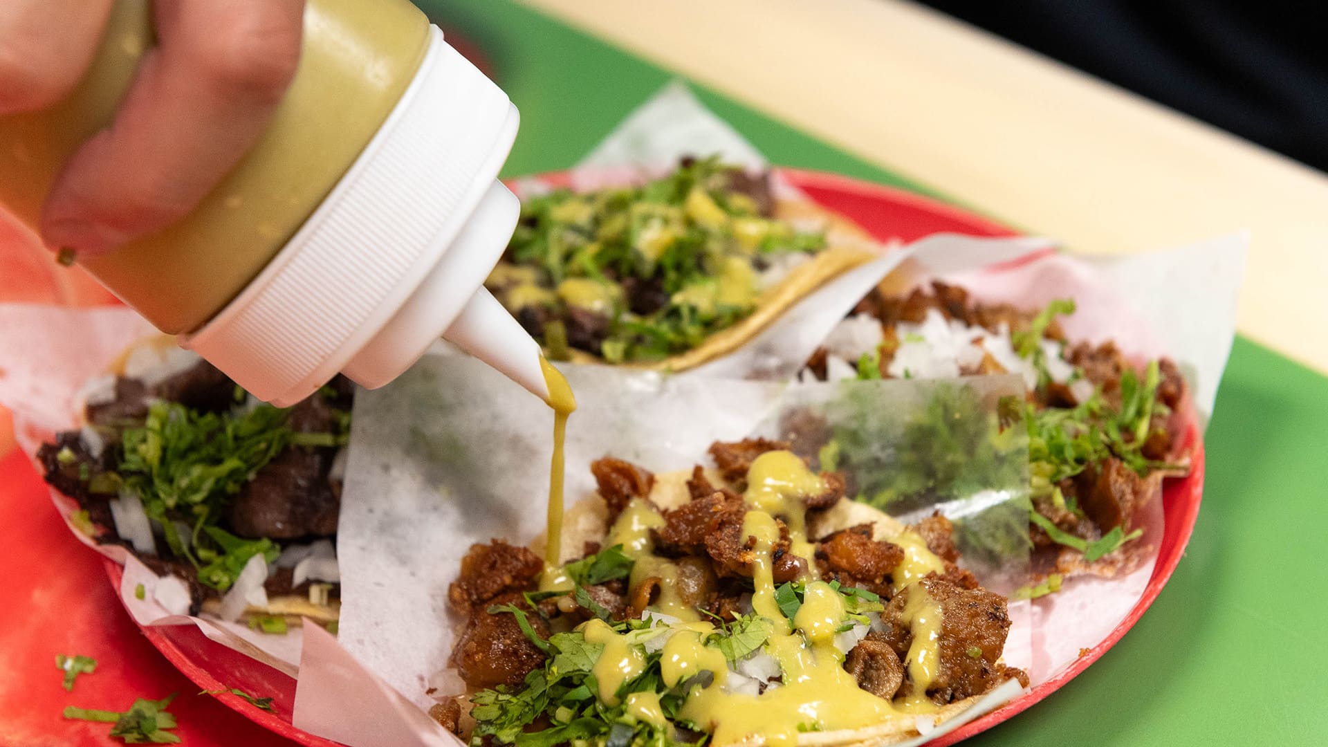 A hand squeezes green salsa over tacos from a bottle