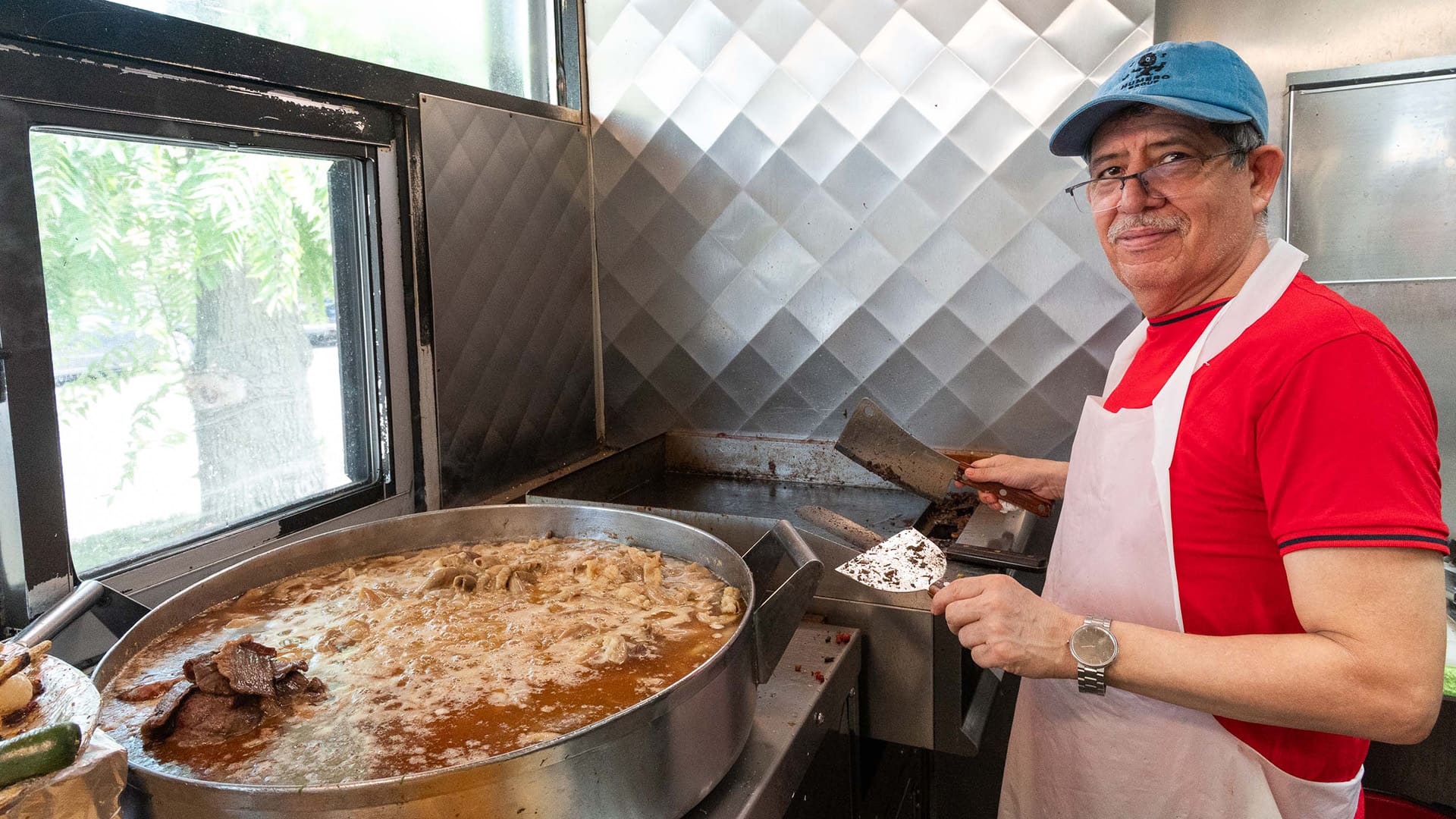 A man in an apron and cap holds a spatula while standing by a big boiling pot