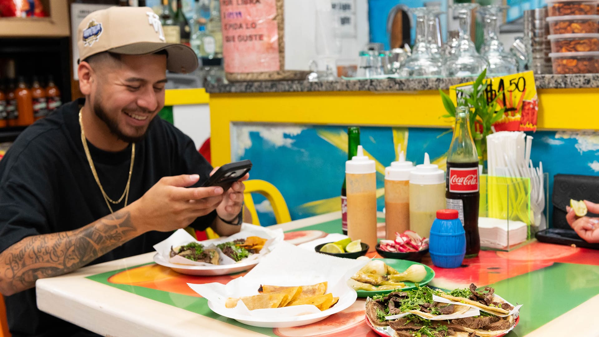 A man in a cap smiles while sitting at a table with two plates of tacos on his phone