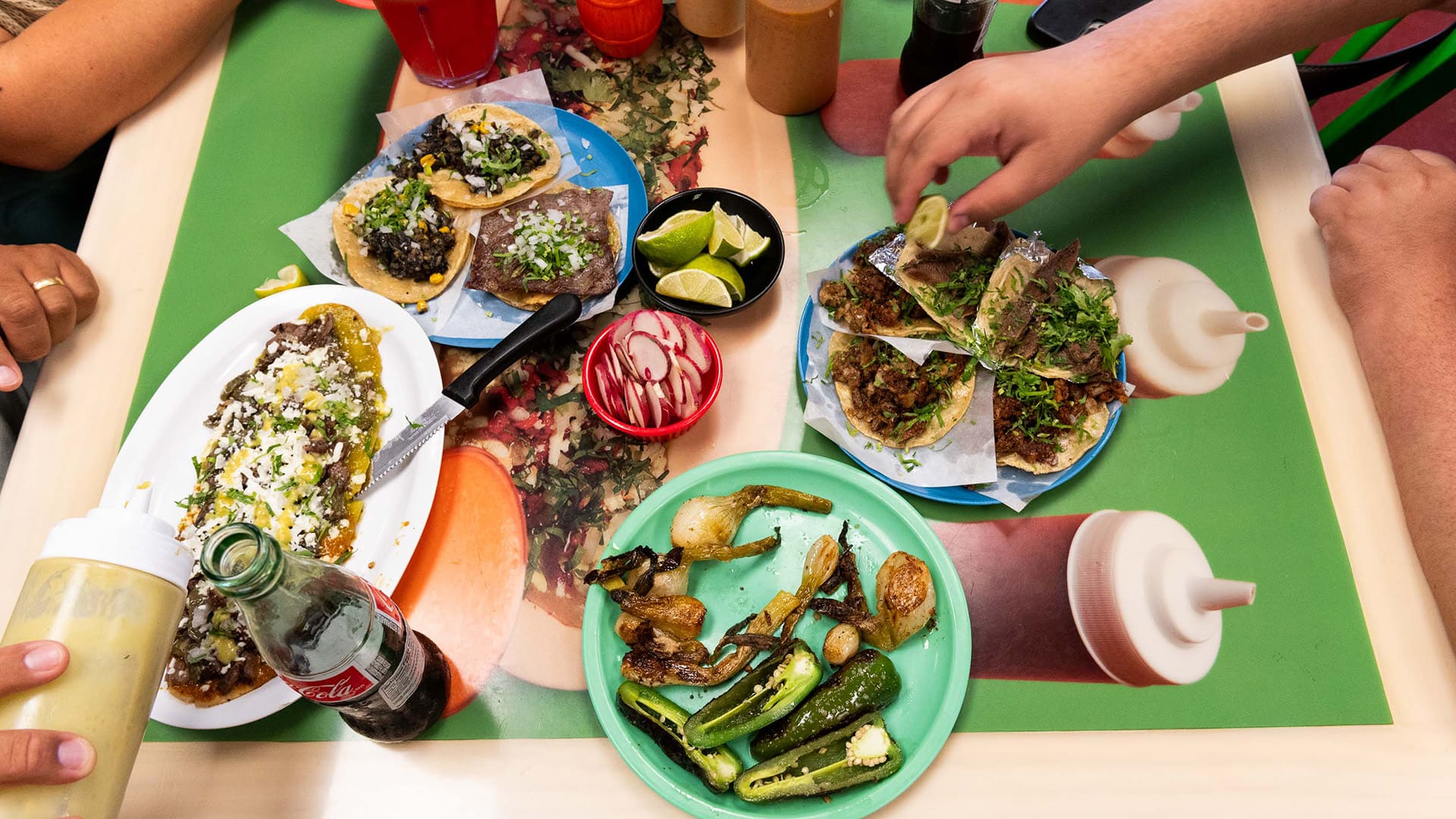 Hands reach over a table full of plates of tacos and a huarache
