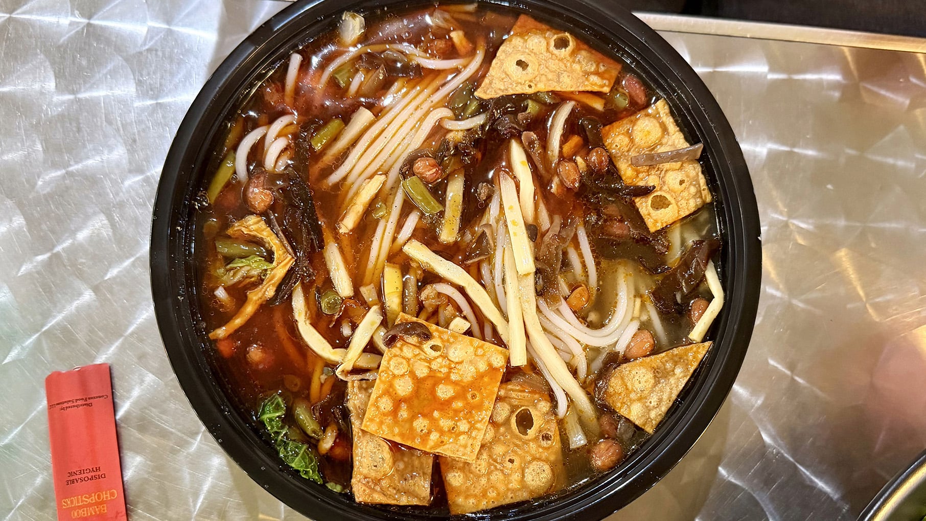 A takeout bowl of noodles and fried tofu skin in a broth